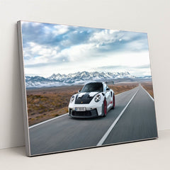 This photo shows a white Porsche 911 GT3 RS with red wheels driving quickly on an open desert highway with mountains in the background, in a silver frame.