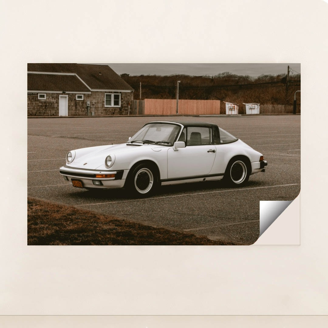 This photo shows a white classic Porsche 911 Targa parked in an empty lot on an overcast day, minimalist unframed style.