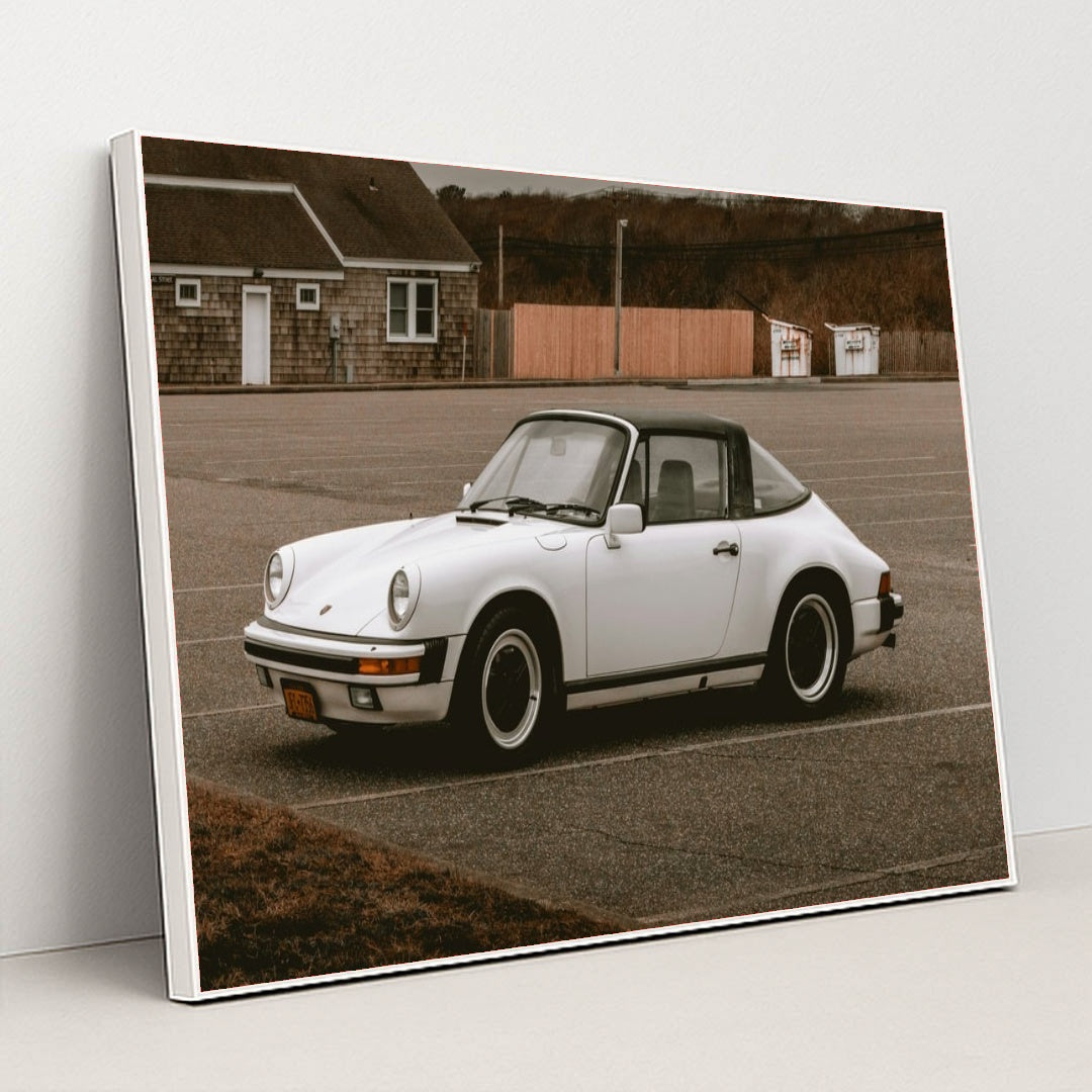 This photo shows a white classic Porsche 911 Targa parked in an empty lot on an overcast day, in a white frame.