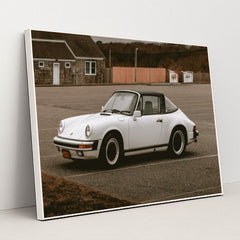 This photo shows a white classic Porsche 911 Targa parked in an empty lot on an overcast day, in a white frame.