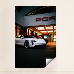 This photo shows a white Porsche Taycan electric sedan parked in front of a Porsche dealership at night, minimalist unframed style.