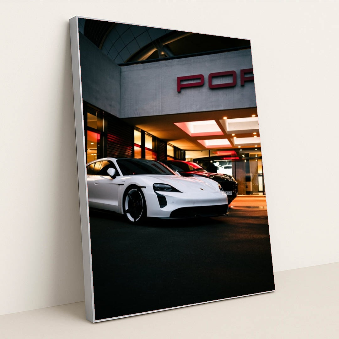 This photo shows a white Porsche Taycan electric sedan parked in front of a Porsche dealership at night, in a silver frame.
