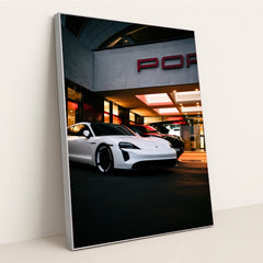 This photo shows a white Porsche Taycan electric sedan parked in front of a Porsche dealership at night, in a silver frame.