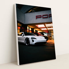This photo shows a white Porsche Taycan electric sedan parked in front of a Porsche dealership at night, in a white frame.