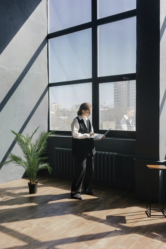 Mikhail Nilov - A person standing by large windows in a modern office, working on a laptop.