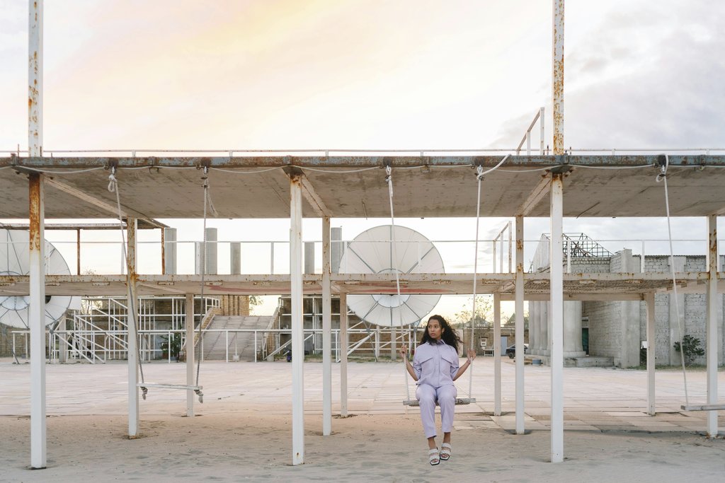 Mikhail Nilov - A woman sits alone on a swing in a deserted industrial area at twilight, evoking solitude.