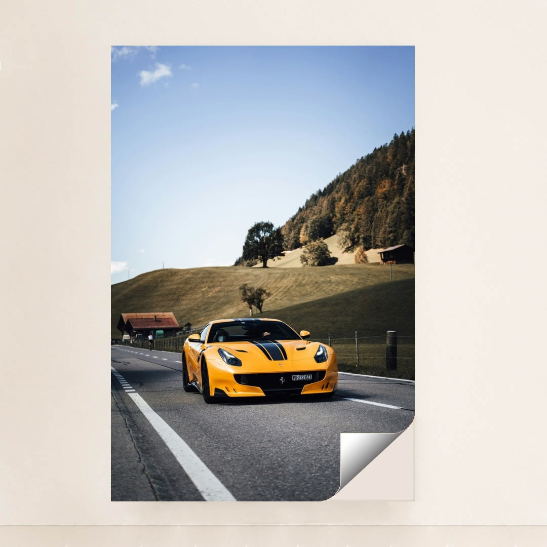 This photo shows a yellow Ferrari sports car with black stripes driving on a rural road by green hills and cottages, minimalist unframed style.