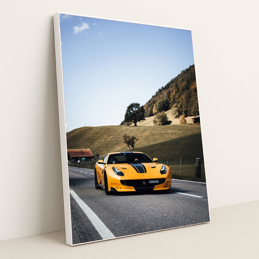 This photo shows a yellow Ferrari sports car with black stripes driving on a rural road by green hills and cottages, in a white frame.