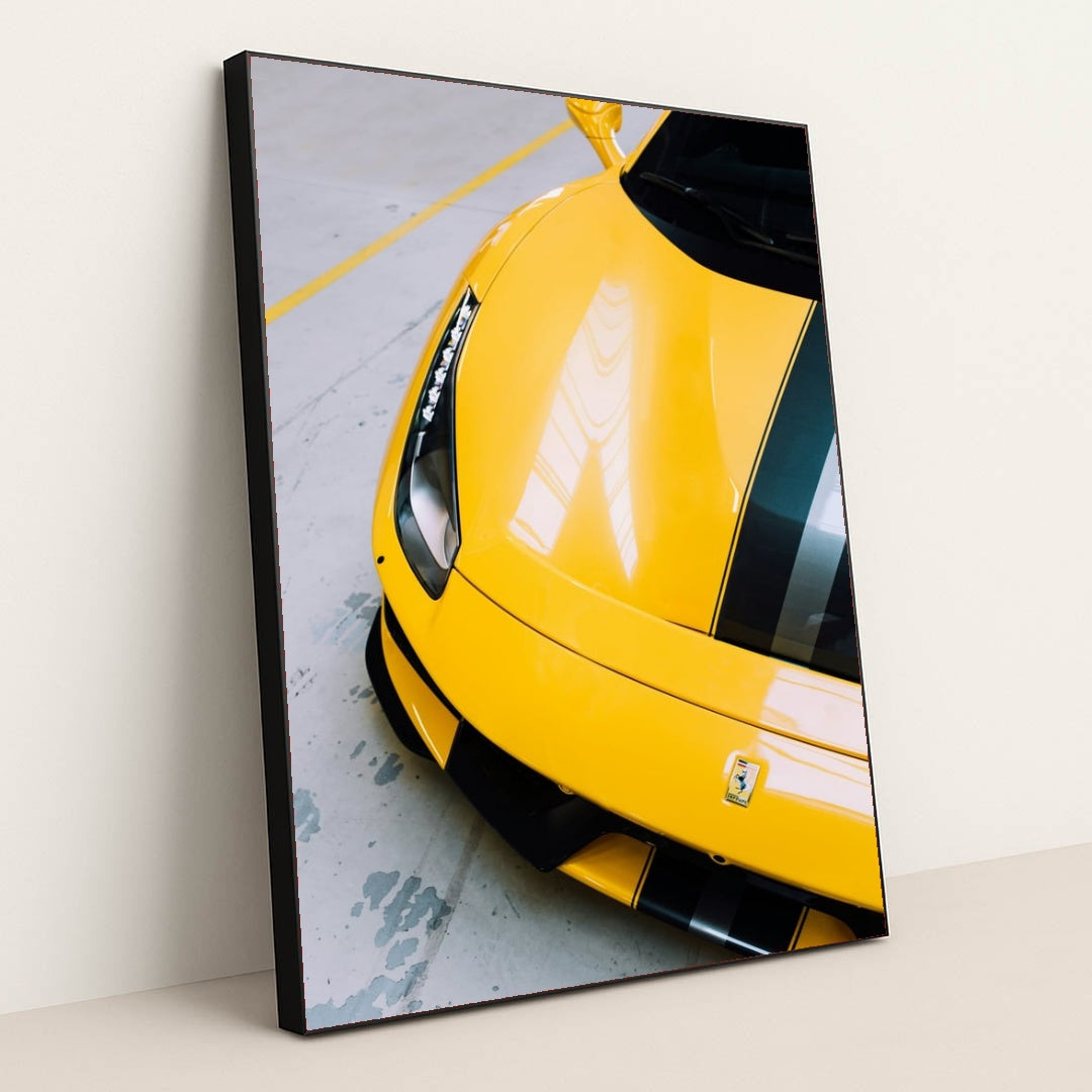 This photo shows the front end of a yellow Ferrari 488 Pista with black and silver racing stripes indoors, in a black frame.