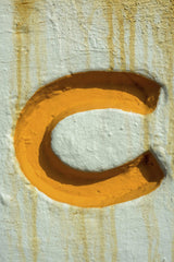 Victor Moragriega - Close-up of an orange letter C painted on a textured wall with rust drip marks.