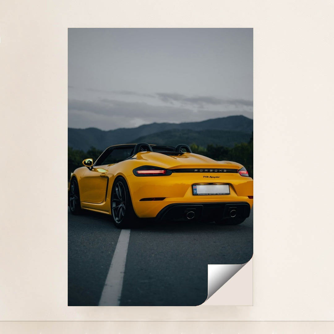 This photo shows the rear of a yellow Porsche 718 Spyder convertible parked on a road with mountains in the background, minimalist unframed style.
