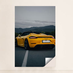 This photo shows the rear of a yellow Porsche 718 Spyder convertible parked on a road with mountains in the background, minimalist unframed style.