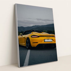 This photo shows the rear of a yellow Porsche 718 Spyder convertible parked on a road with mountains in the background, in a silver frame.