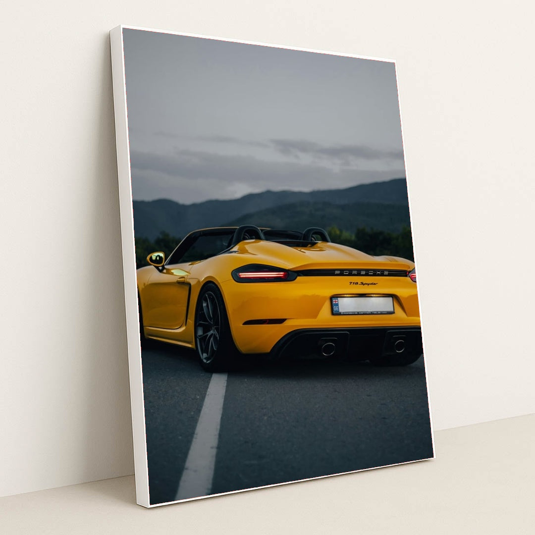 This photo shows the rear of a yellow Porsche 718 Spyder convertible parked on a road with mountains in the background, in a white frame.