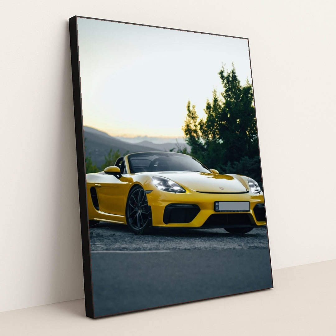 This photo shows a yellow Porsche convertible sports car parked at sunset near green trees and mountains, in a black frame.