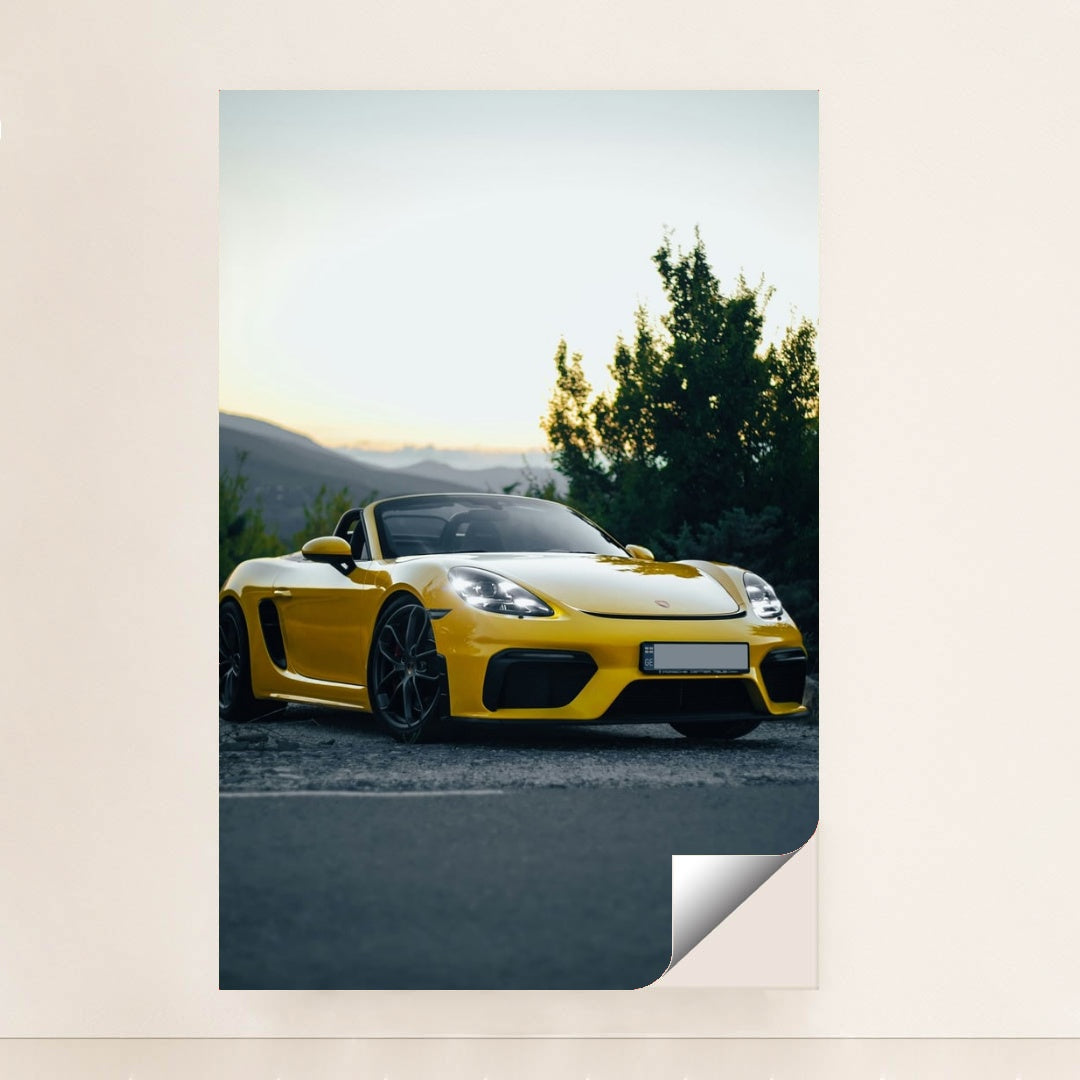 This photo shows a yellow Porsche convertible sports car parked at sunset near green trees and mountains, minimalist unframed style.