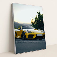 This photo shows a yellow Porsche convertible sports car parked at sunset near green trees and mountains, in a silver frame.