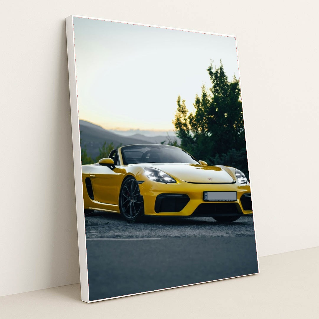 This photo shows a yellow Porsche convertible sports car parked at sunset near green trees and mountains, in a white frame.