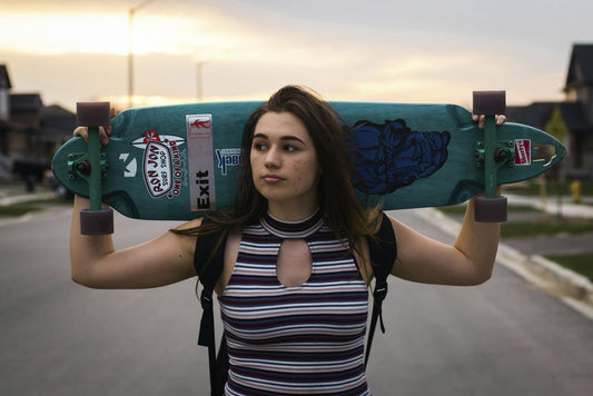 Trinity  Kubassek - Teen girl carries a longboard skateboard on shoulders in suburban street at sunset, showcasing modern skate style.