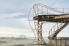 Mikhail Nilov - An abandoned rusty staircase structure on a deserted beach with a cloudy sky.