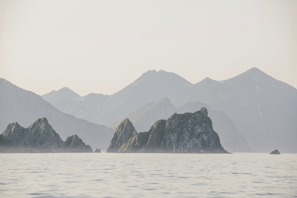 Julia Volk - A tranquil view of rocky coastal mountains reflecting on calm water at dusk, surrounded by a pastel sky.