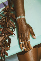 Jess Loiterton - Close-up of adorned hand holding a surfboard, highlighting jewelry against a vibrant beach backdrop.