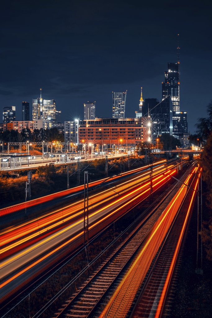Adam Borkowski - Dynamic night view of Warsaw skyline with vibrant train light trails creating motion effect.