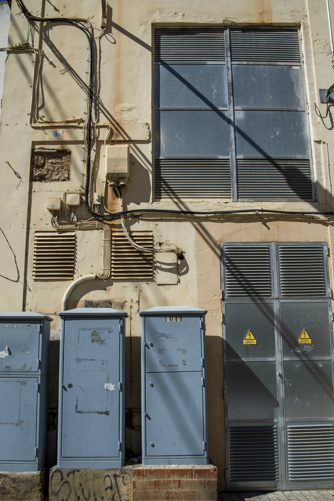 Victor  Moragriega - Urban industrial building facade with blue electrical boxes and cables in sunlight.