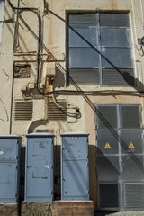 Victor  Moragriega - Urban industrial building facade with blue electrical boxes and cables in sunlight.
