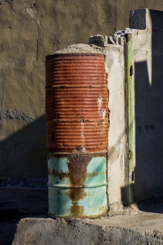 Victor  Moragriega - A rusty metal barrel standing next to a weathered wall, showcasing decay and urban textures.