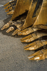 Victor  Moragriega - Detailed view of worn rusted excavator buckets lined up on rough concrete.