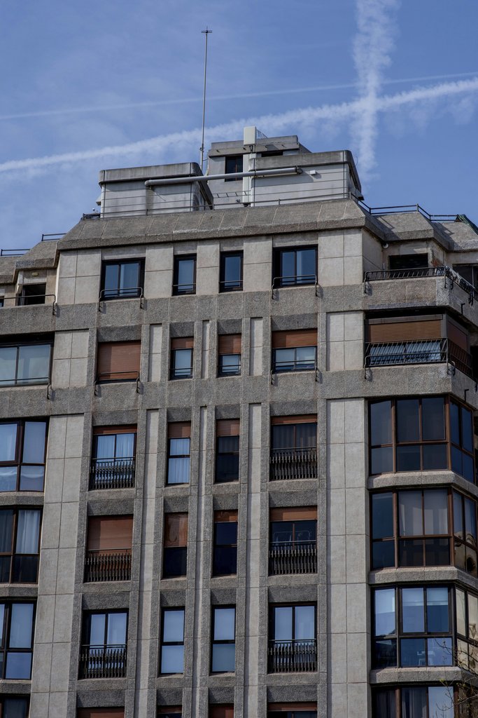 Victor  Moragriega - Concrete high-rise apartment building under clear blue sky with contrails, city architecture.