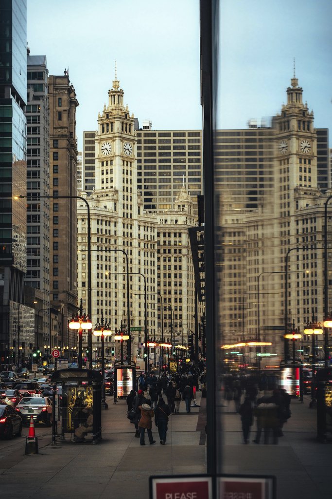 Mike Norris - Vibrant street scene of Chicago's Wrigley Building reflected on glass facade, evening rush hour.