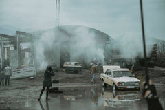 Erik Uruci - Vintage Mercedes car in a smoky industrial area with people and puddle reflections.