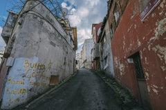 Sami TÜRK - A narrow, untidy urban alleyway featuring weathered buildings and graffiti under a cloudy sky.