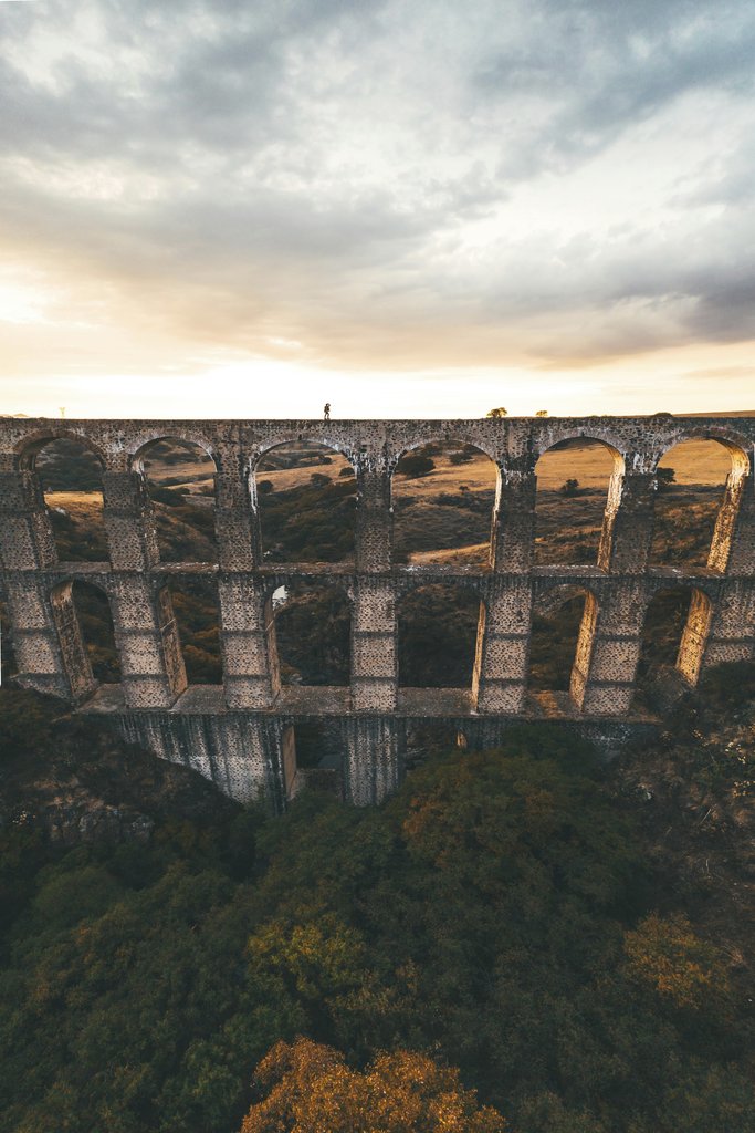 Mikhail Nilov - A breathtaking aerial shot of an ancient aqueduct amidst lush greenery during sunset.