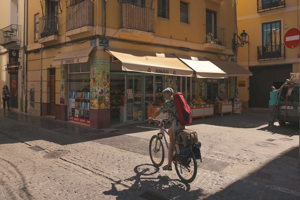 TBD Traveller - A cyclist rides past a shop in a sunny street corner in Barcelona, capturing local urban life.