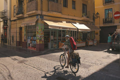 TBD Traveller - A cyclist rides past a shop in a sunny street corner in Barcelona, capturing local urban life.
