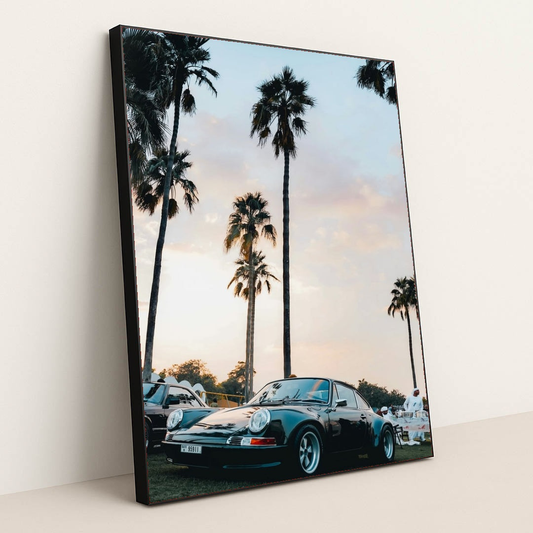 This photo shows a classic black Porsche sports car parked under tall palm trees at sunset, in a black frame.