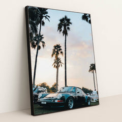 This photo shows a classic black Porsche sports car parked under tall palm trees at sunset, in a black frame.