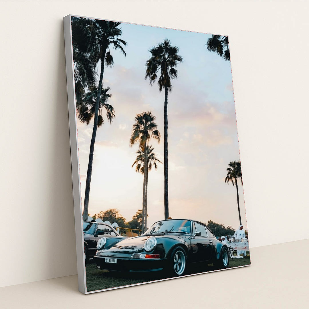 This photo shows a classic black Porsche sports car parked under tall palm trees at sunset, in a silver frame.