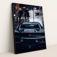 This photo shows the rear view of a black Ferrari covered in rain droplets on a wet city street at night, in a black frame.