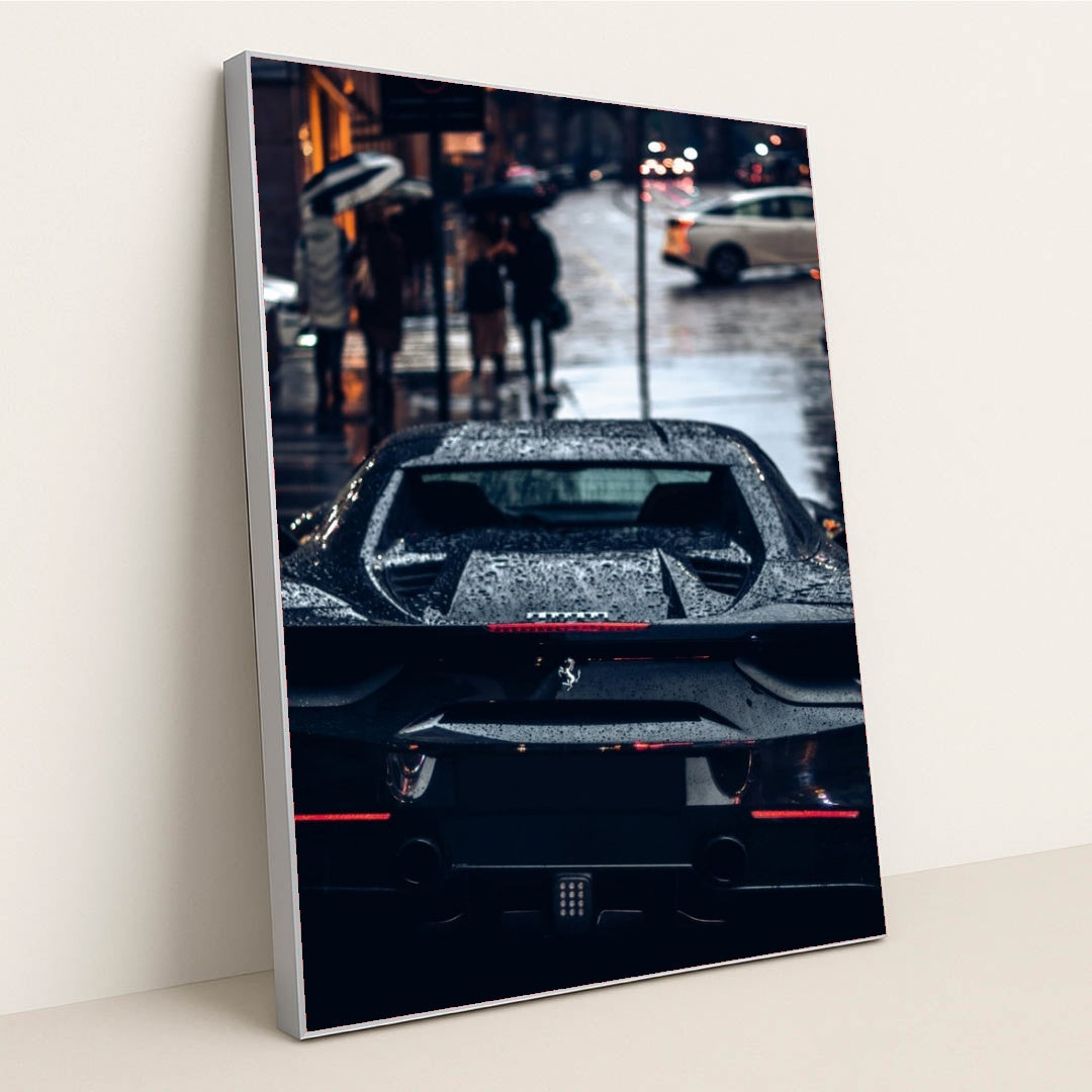 This photo shows the rear view of a black Ferrari covered in rain droplets on a wet city street at night, in a silver frame.