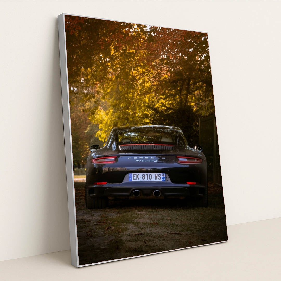This photo shows the rear view of a black Porsche 911 Carrera S parked under autumn trees in soft evening light, in a silver frame.