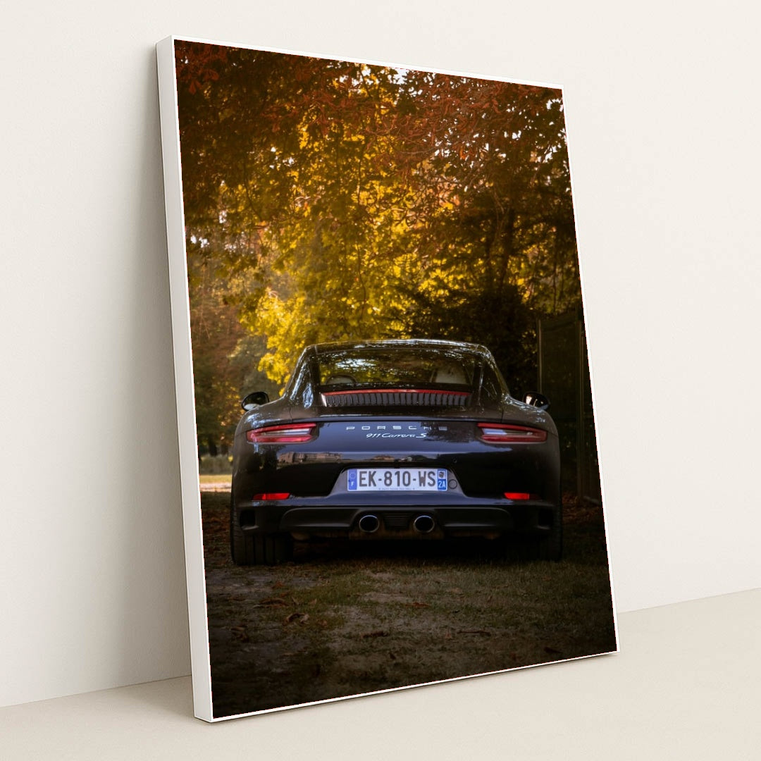 This photo shows the rear view of a black Porsche 911 Carrera S parked under autumn trees in soft evening light, in a white frame.