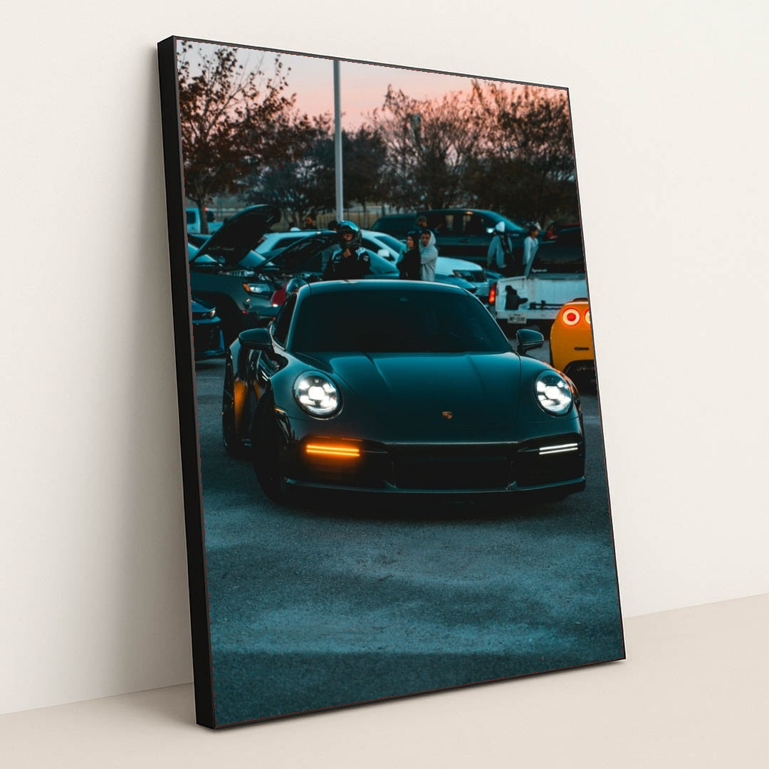This photo shows a black Porsche 911 with LED headlights at dusk, parked at a car meet with people and vehicles in the background, in a black frame.