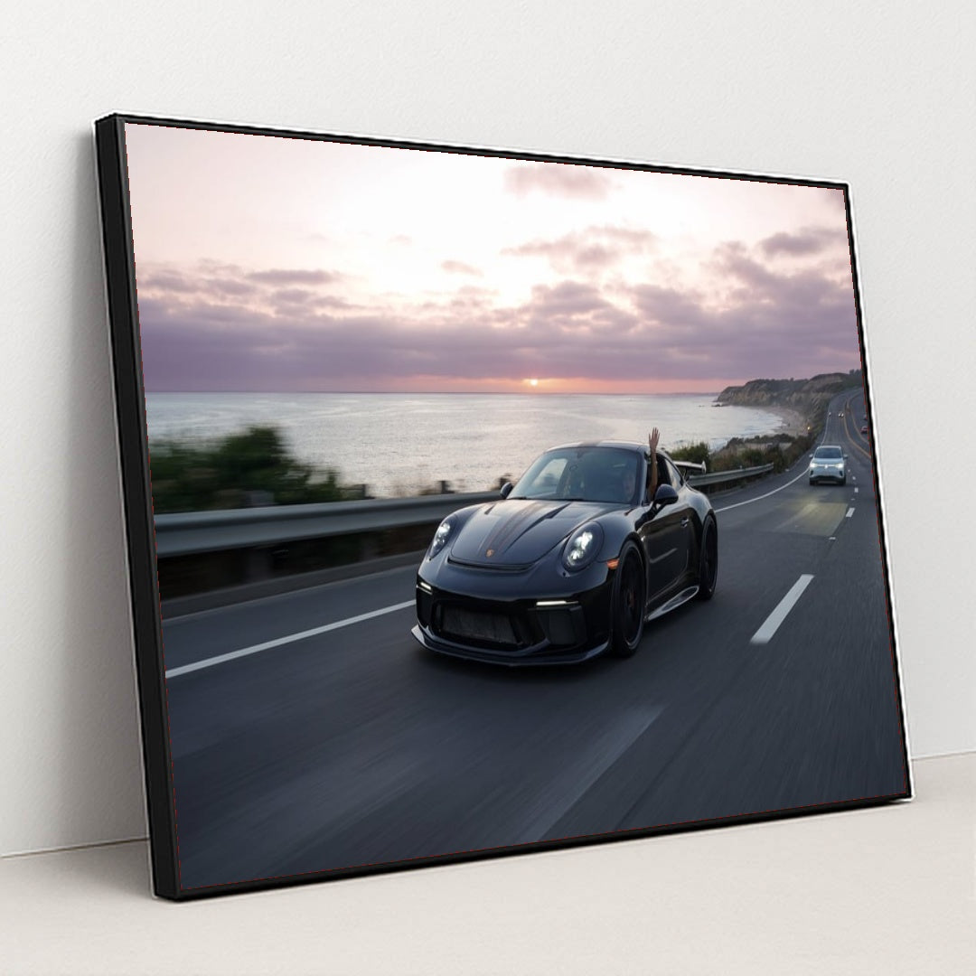 This photo shows a black Porsche speeding down a coastal highway at sunset with a hand waving from the window, in a black frame.