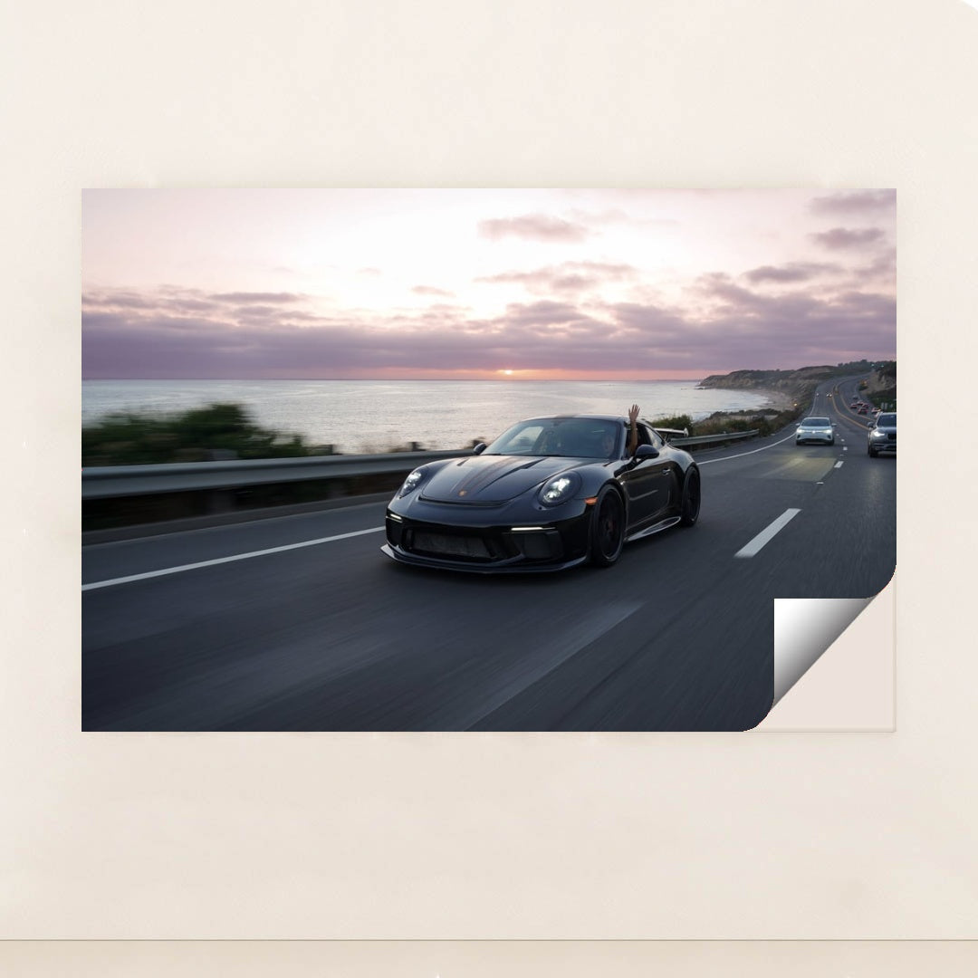 This photo shows a black Porsche speeding down a coastal highway at sunset with a hand waving from the window, unframed.