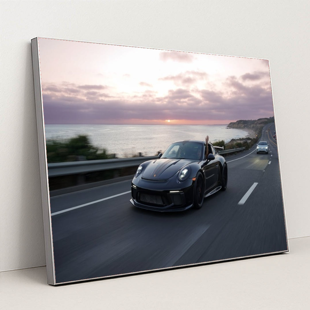 This photo shows a black Porsche speeding down a coastal highway at sunset with a hand waving from the window, in a silver frame.