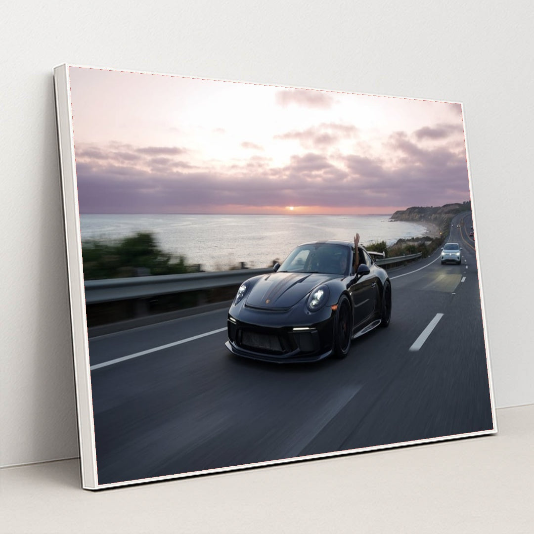 This photo shows a black Porsche speeding down a coastal highway at sunset with a hand waving from the window, in a white frame.
